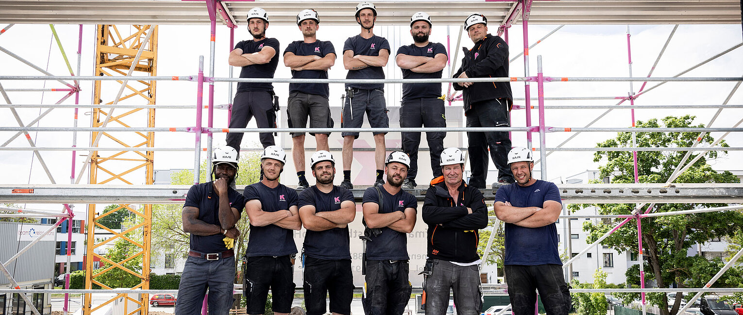 Zehn Bauarbeiter in Arbeitskleidung und Schutzhelmen posieren auf einer Baustelle vor einem Gerüst, im Hintergrund sind Baukräne und Gebäude zu sehen.