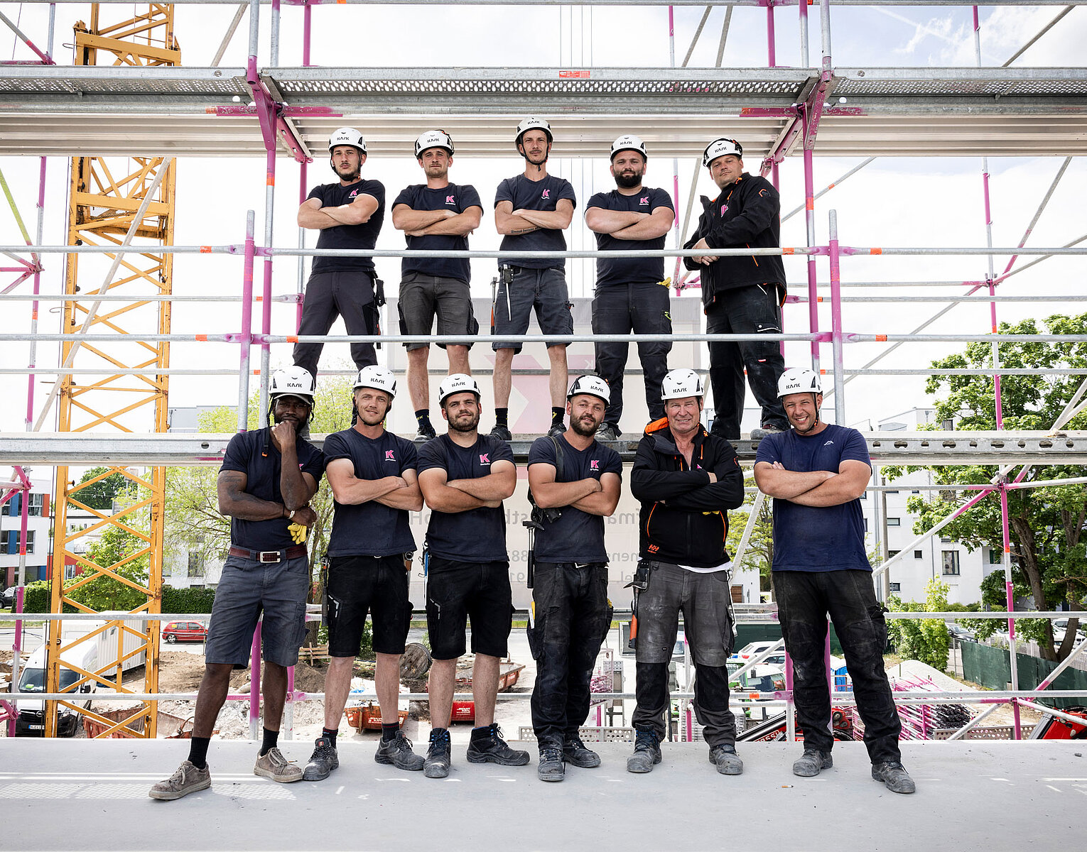 Gerüstbau Kaufmann Zehn Bauarbeiter in Arbeitskleidung und Schutzhelmen posieren auf einer Baustelle vor einem Gerüst, im Hintergrund sind Baukräne und Gebäude zu sehen.
