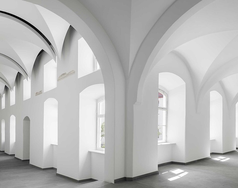 Innenansicht eines weißen Raums mit hohen gewölbten Decken, großen Fenstern und weißem Fußboden, durch das Tageslicht gut beleuchtet.