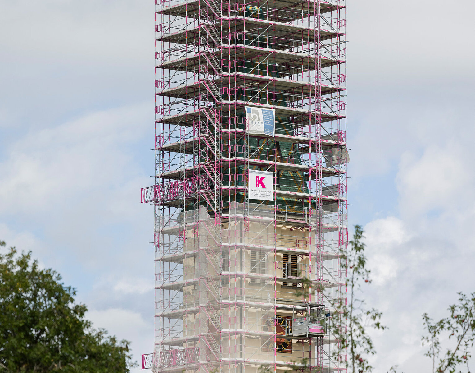 Kirchturm untergerüstet mit einem Gerüst aus Metallrohren, umgeben von Bäumen und bewölktem Himmel.