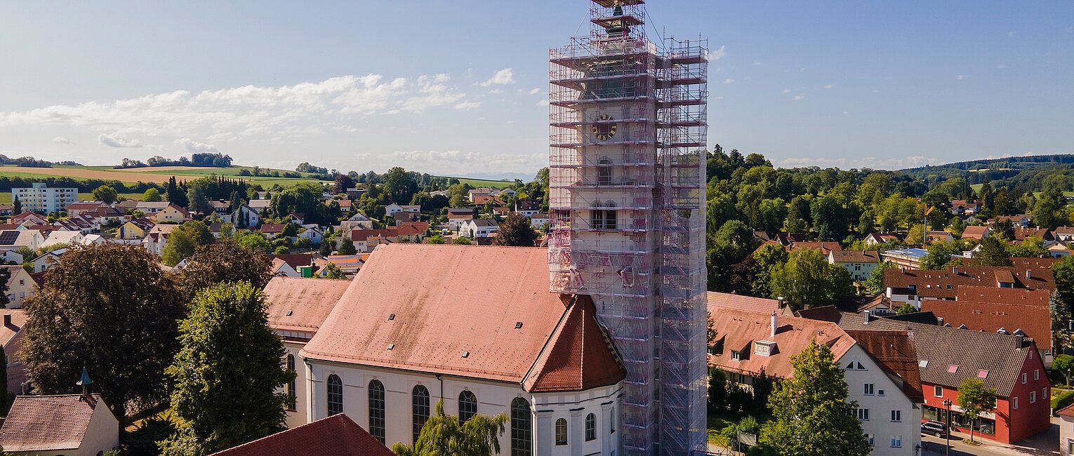 Kirchturm eines historischen Gebäudes ist von Gerüsten umgeben, umgeben von Häusern und viel Grün unter blauem Himmel.