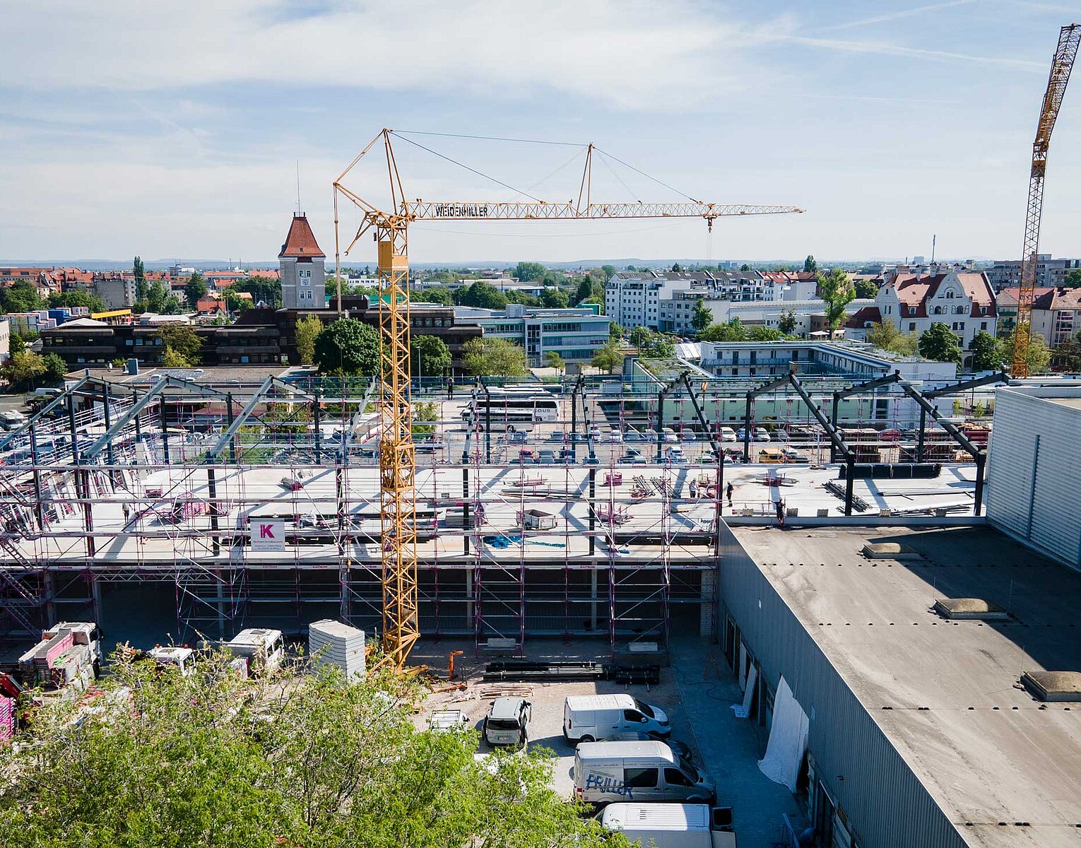 Baustelle mit einem großen Kran, Stahlgerüsten und mehreren Fahrzeugen, umgeben von Gebäuden und Vegetation in einer Stadtlandschaft bei klarem Himmel.