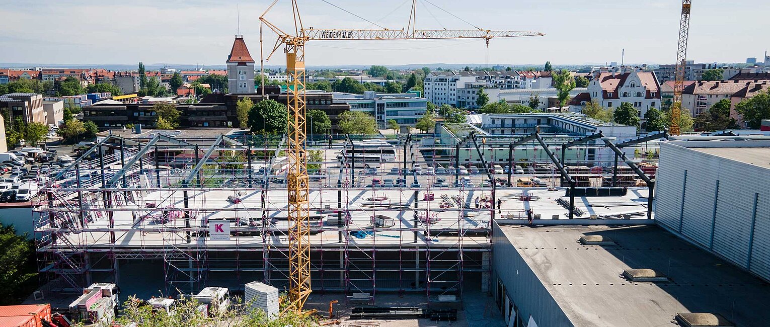 Baustelle mit einem großen Kran, Stahlgerüsten und mehreren Fahrzeugen, umgeben von Gebäuden und Vegetation in einer Stadtlandschaft bei klarem Himmel.