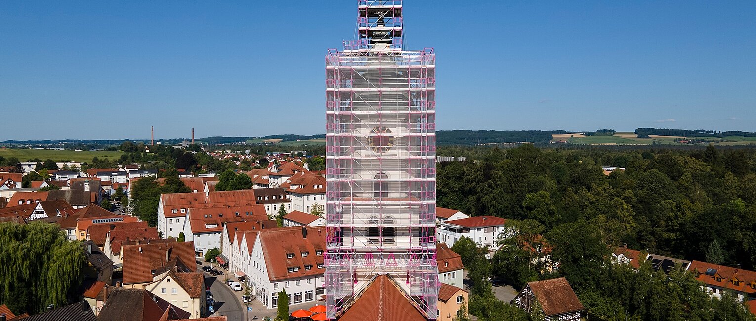 Stadtsilhouette mit einem Turm im Zentrum, der von einem Gerüst umgeben ist, umgeben von roten Dächern und grünen Bäumen unter klarem blauem Himmel.
