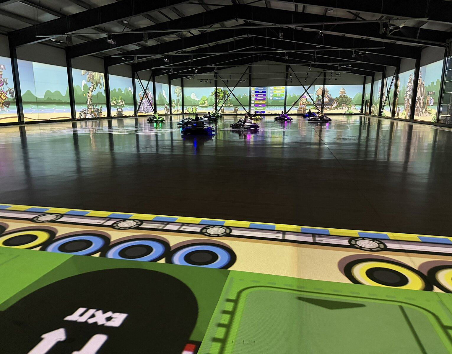 Innenansicht einer Halle mit mehreren fahrbaren Kartfahrzeugen auf einer glatten Fläche, umgeben von bunten, spielerischen Wandprojektionen.