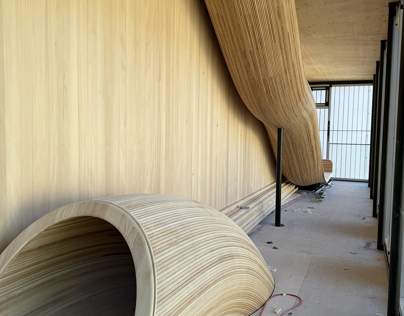 Flur mit zwei großen, geschwungenen, holzverkleideten Sitz- oder Schaukelelementen, einer an der Wand montiert und einer auf dem Boden stehend, umgeben von Holzverkleidungen und Baustellenmaterialien.