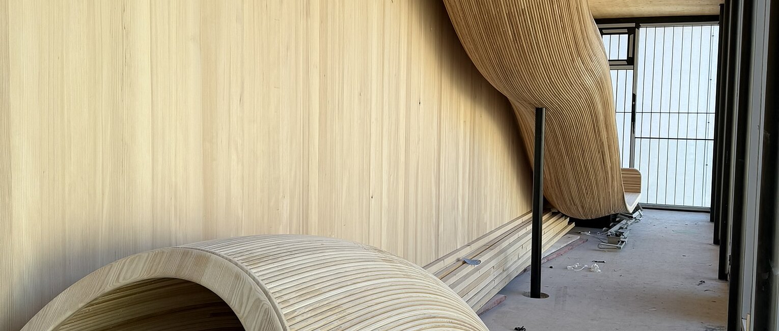 Flur mit zwei großen, geschwungenen, holzverkleideten Sitz- oder Schaukelelementen, einer an der Wand montiert und einer auf dem Boden stehend, umgeben von Holzverkleidungen und Baustellenmaterialien.