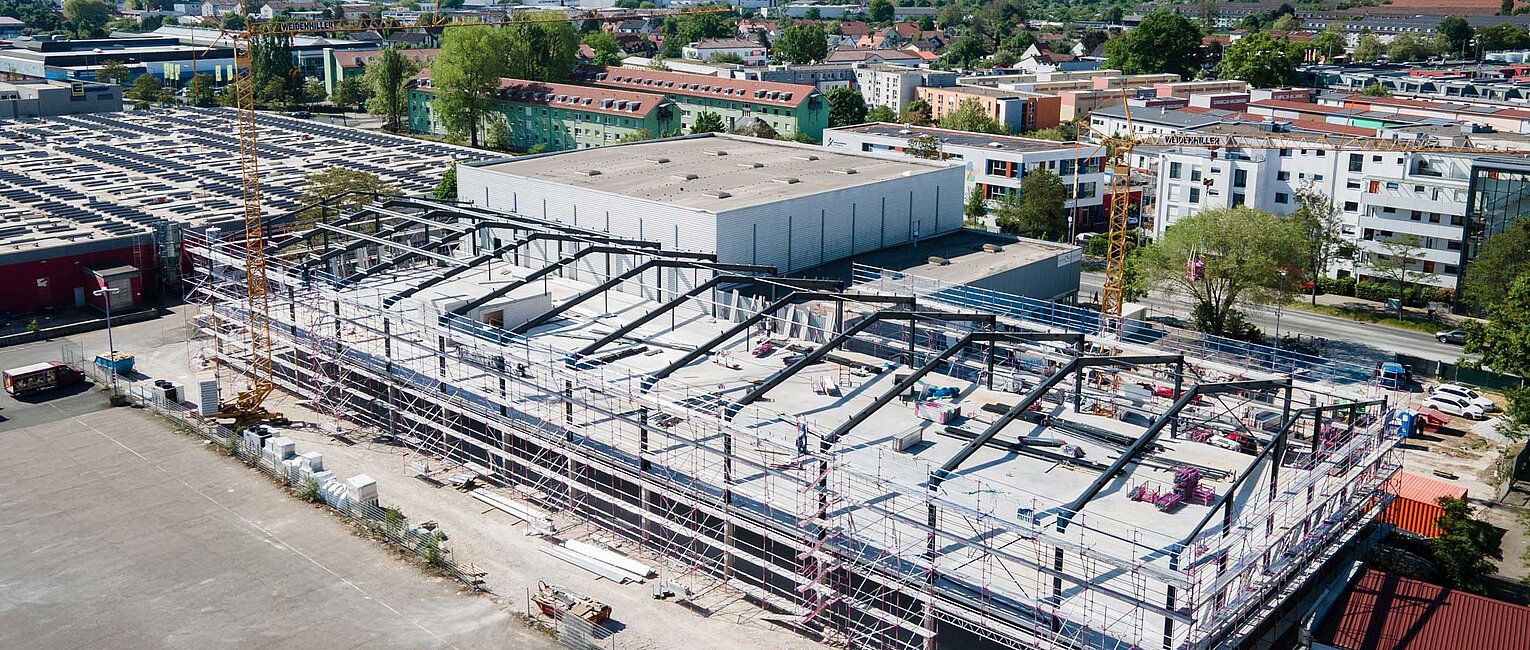 Luftaufnahme einer großen Baustelle eines Industriegebäudes mit Stahlrahmen, umgeben von Wohn- und Gewerbegebäuden in einer Stadt.