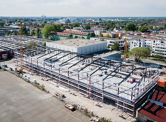 Luftaufnahme einer großen Baustelle eines Industriegebäudes mit Stahlrahmen, umgeben von Wohn- und Gewerbegebäuden in einer Stadt.