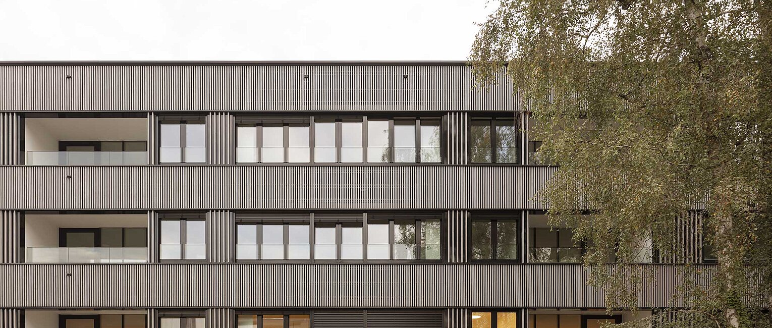 Mehrstöckiges modernes Wohngebäude mit großen Fenstern und Balkonen, daneben ein Baum mit grünen Blättern.