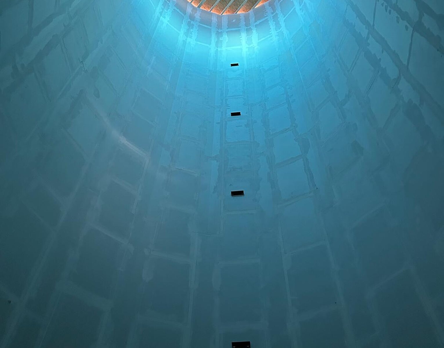 Blick nach oben in einen hohen, zylindrischen Schacht mit einer Gitterstruktur am oberen Ende und blauem Licht, das den Schacht beleuchtet.