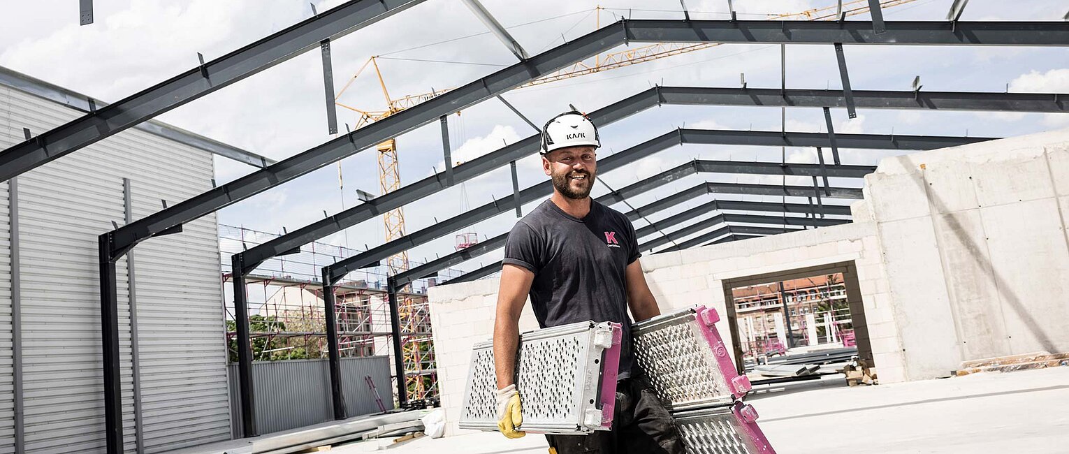 Bauarbeiter mit Helm trägt zwei Metallplatten auf einer Baustelle in einem unvollendeten Stahlrahmengebäude bei Tageslicht.