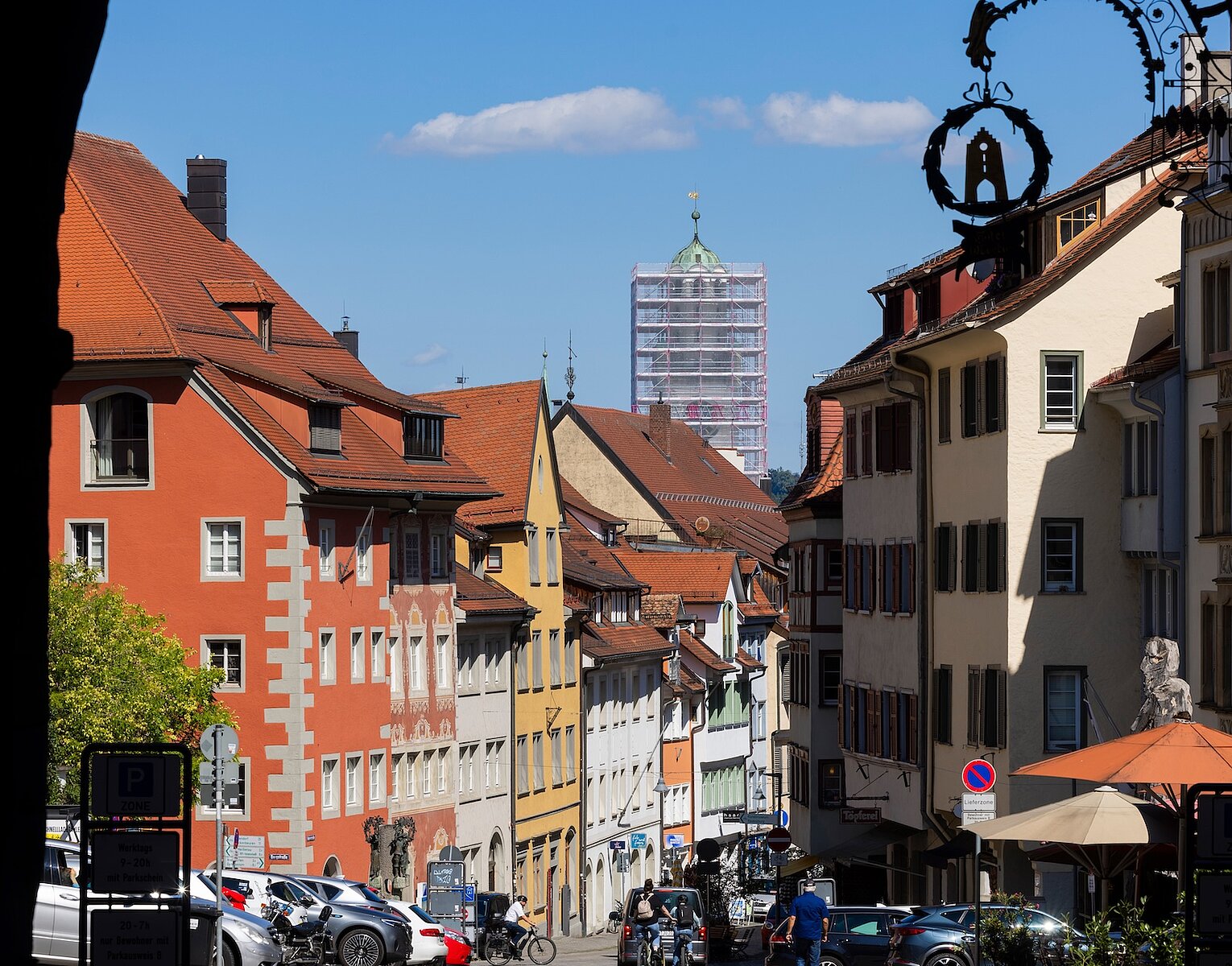 Blick auf eine Altstadtstraße mit bunten historischen Gebäuden, parkenden Autos und Menschen, im Hintergrund ein Kirchturm unter Gerüst.