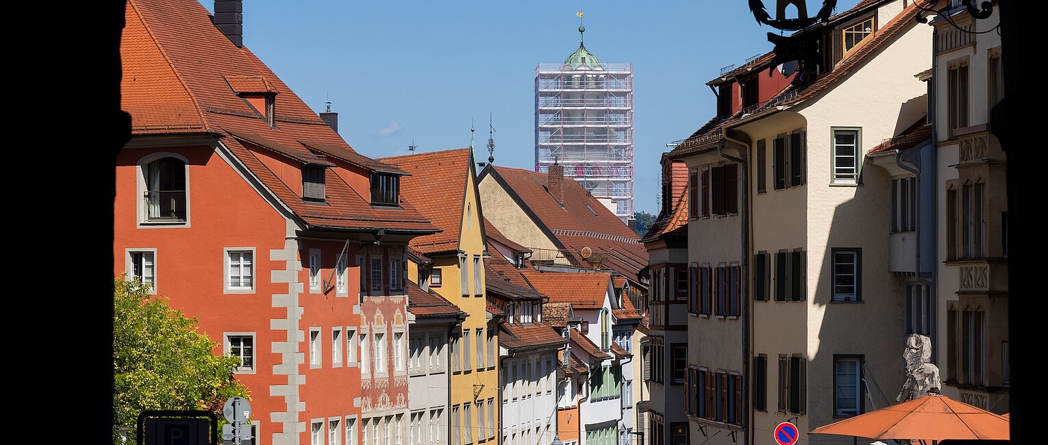 Blick auf eine Altstadtstraße mit bunten historischen Gebäuden, parkenden Autos und Menschen, im Hintergrund ein Kirchturm unter Gerüst.