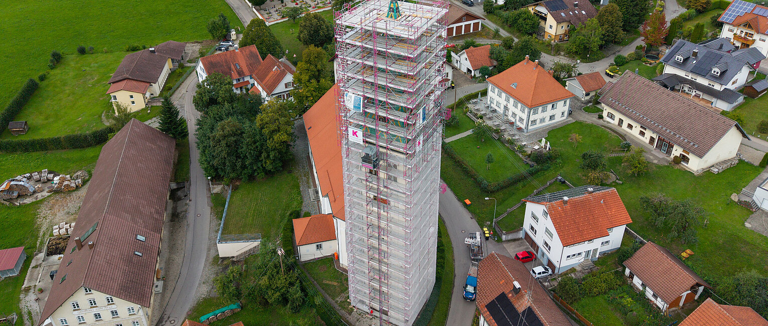 Luftaufnahme eines Dorfes mit einem hohen, eingerüsteten Turm in der Mitte, umgeben von Wohnhäusern, Straßen und grünen Wiesen.