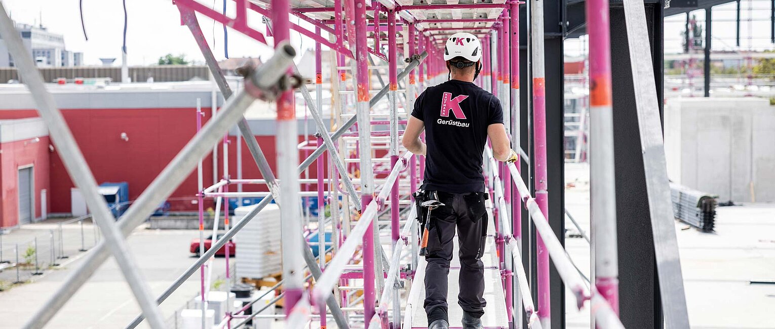 Ein Bauarbeiter mit Schutzhelm und schwarzem T-Shirt geht auf einem Gerüst mit pinkfarbenen Metallstreben. Im Hintergrund sieht man Gebäude und Baustellenausrüstung.