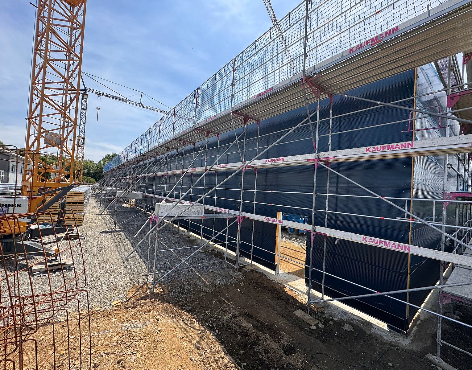 Baustelle mit Gerüst um ein langes Gebäude, Baukran im Hintergrund, sonniger Himmel.