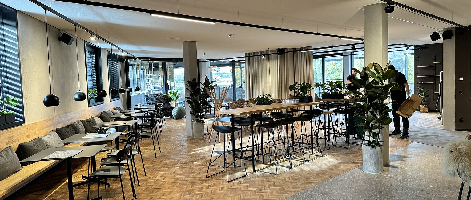 Moderner, heller Gemeinschaftsraum mit Tischen und Stühlen, großen Fenstern, Pflanzen und Holzfußboden in einem Büro oder Café.