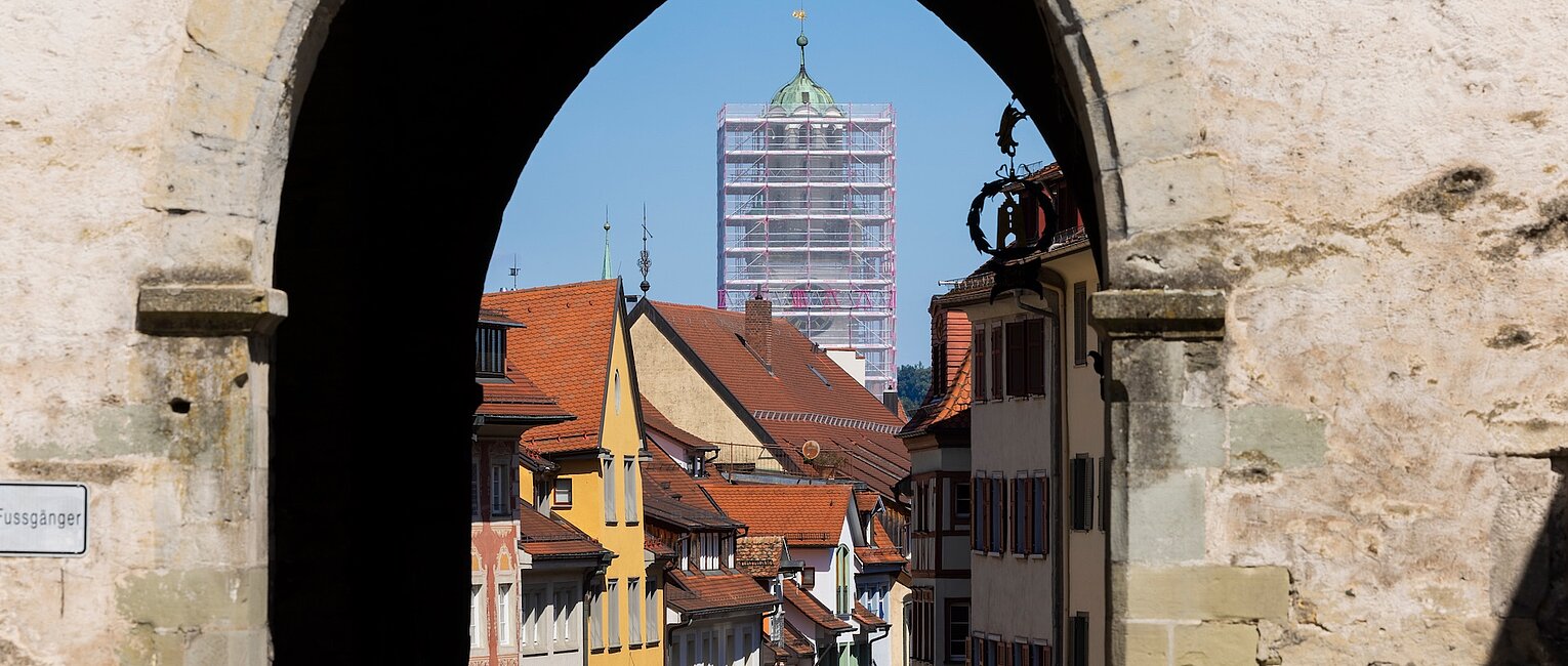 Blick durch einen steinernen Torbogen auf eine Altstadt mit bunten Fachwerkhäusern und einem hohen Kirchturm im Hintergrund, der von Gerüsten umgeben ist.