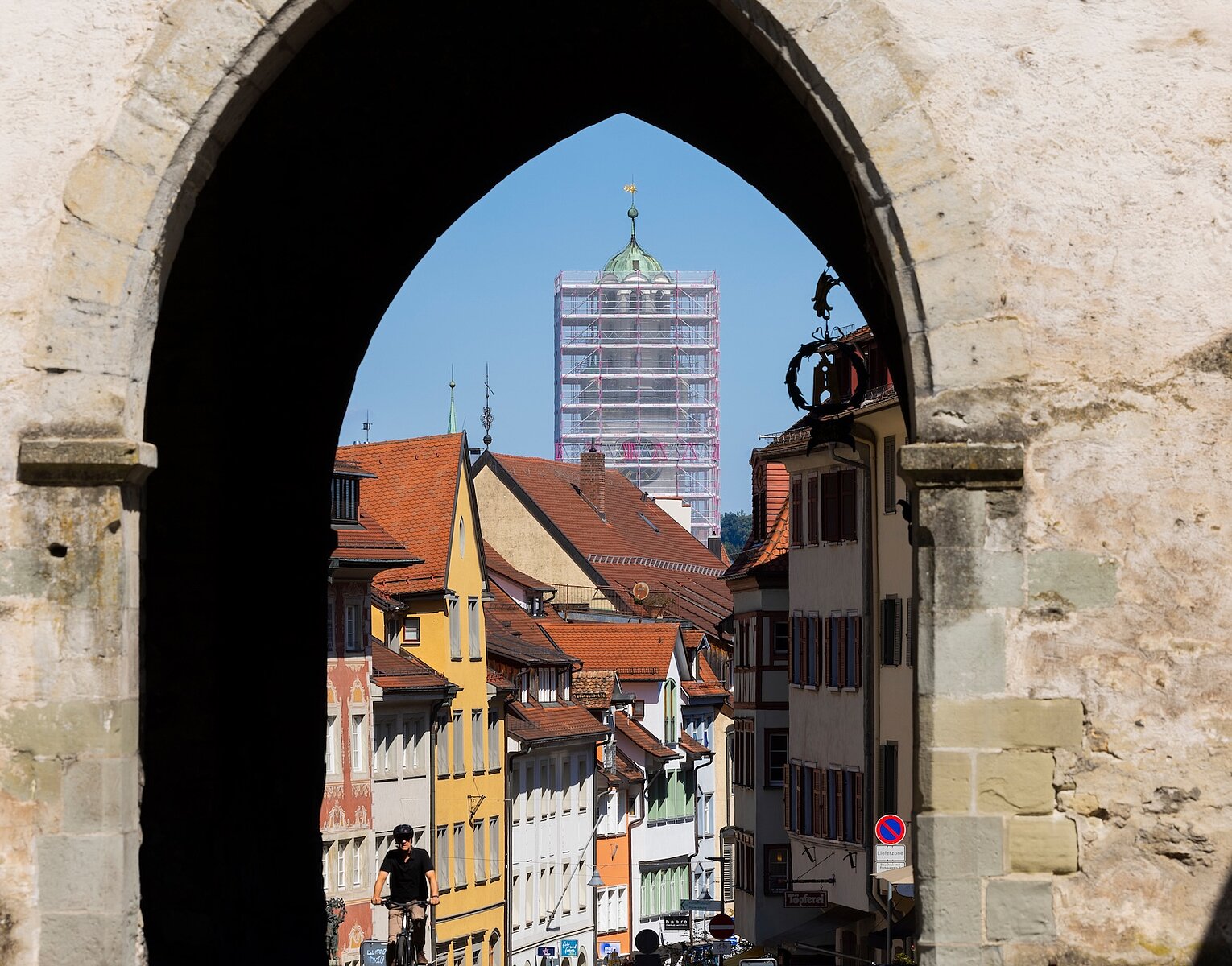 Blick durch einen steinernen Torbogen auf eine Altstadt mit bunten Fachwerkhäusern und einem hohen Kirchturm im Hintergrund, der von Gerüsten umgeben ist.
