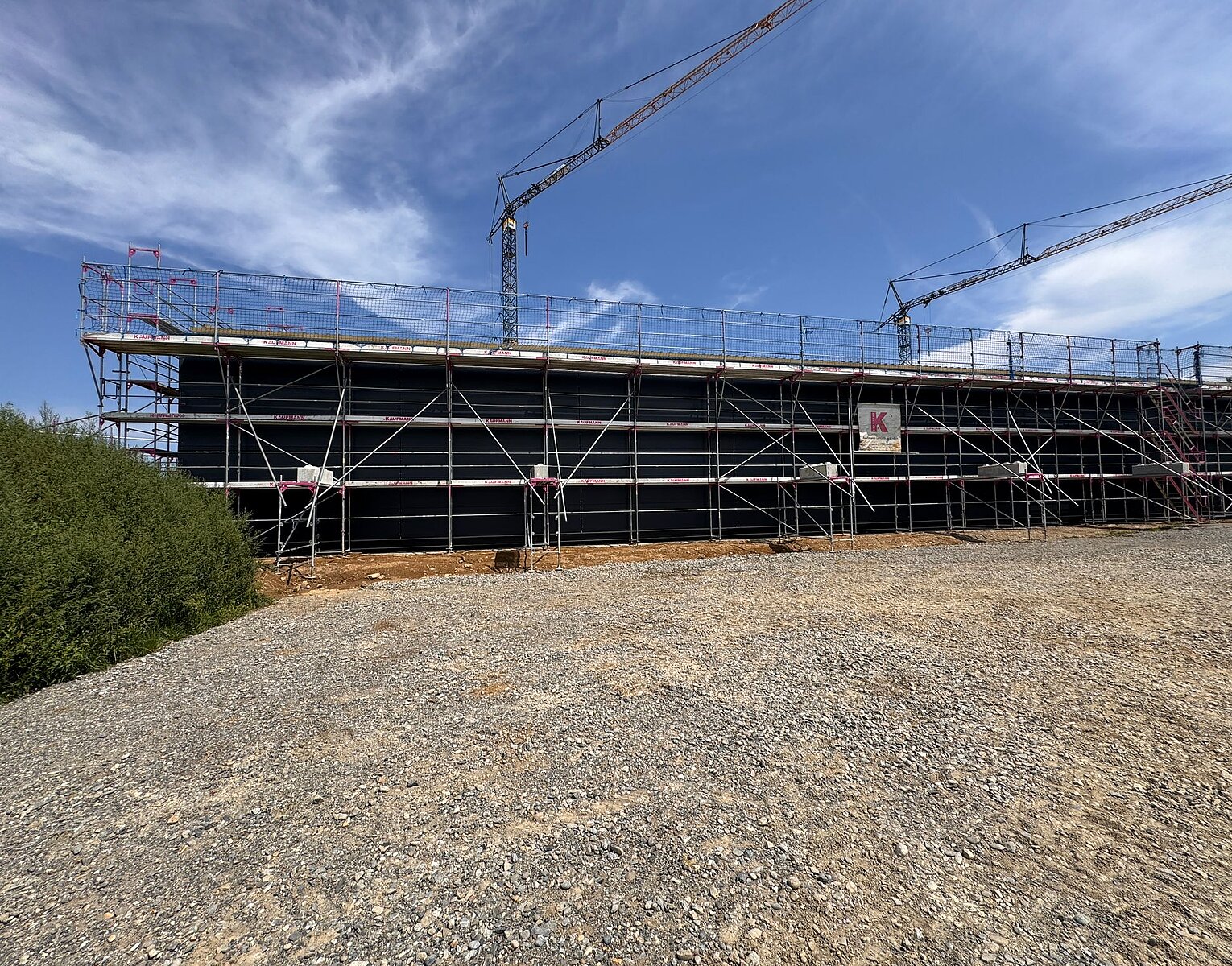 Große Baustelle mit Gerüst um ein rechteckiges Gebäude, zwei großen Kränen und freiem Himmel im Hintergrund.