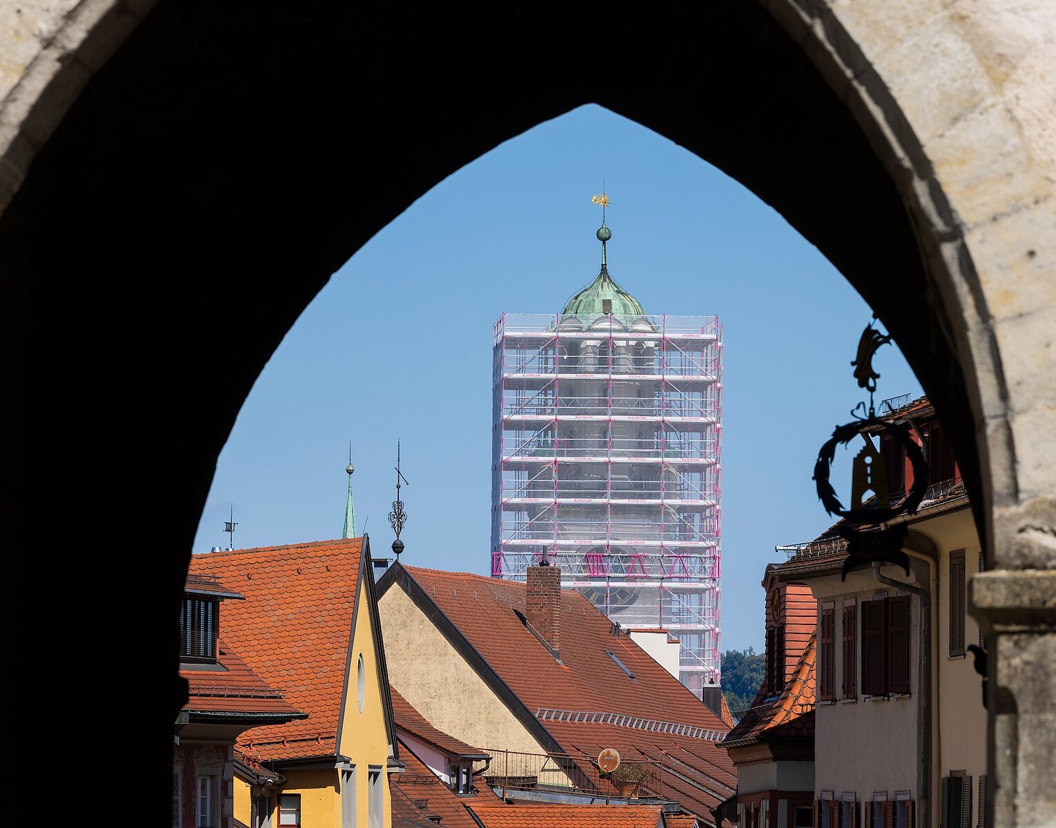 Blick durch einen historischen Torbogen auf ein Turmgerüst und Dächer eines kleinen Ortes bei klarem Himmel.