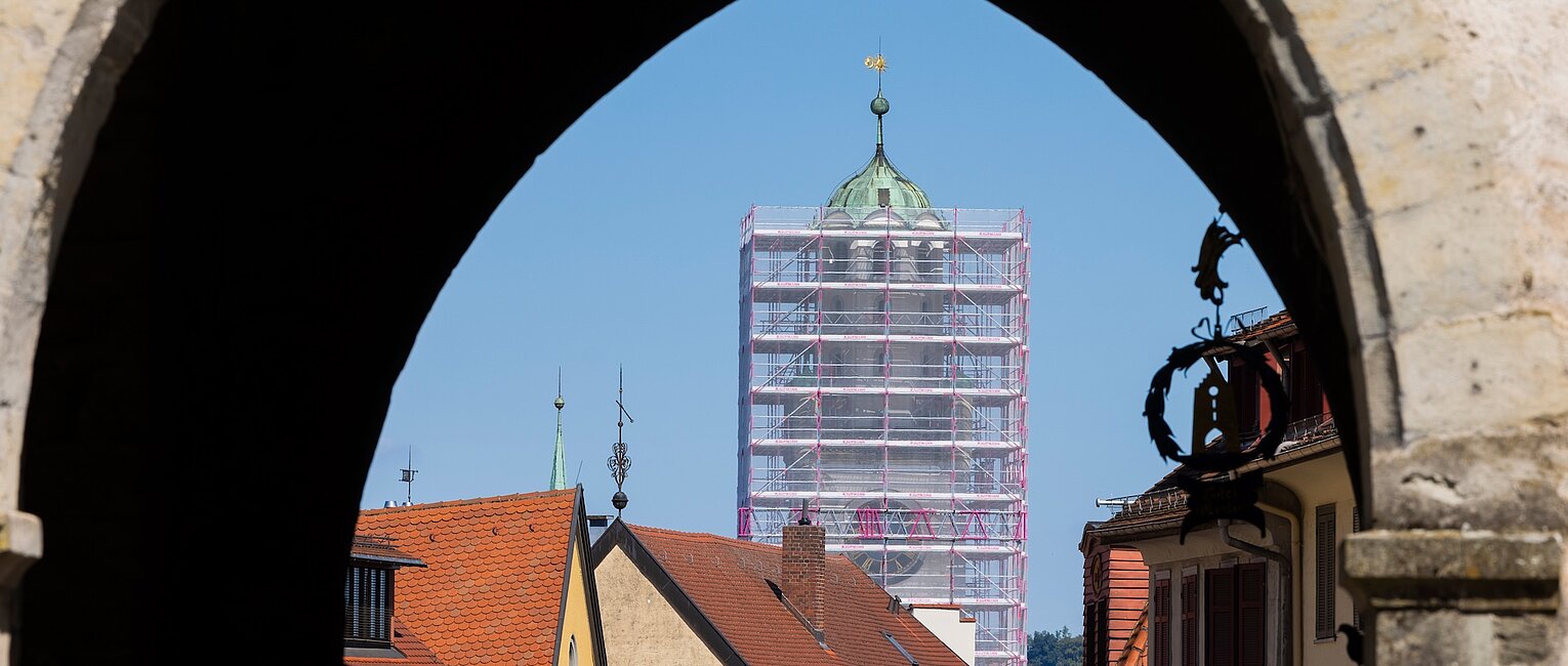 Blick durch einen historischen Torbogen auf ein Turmgerüst und Dächer eines kleinen Ortes bei klarem Himmel.