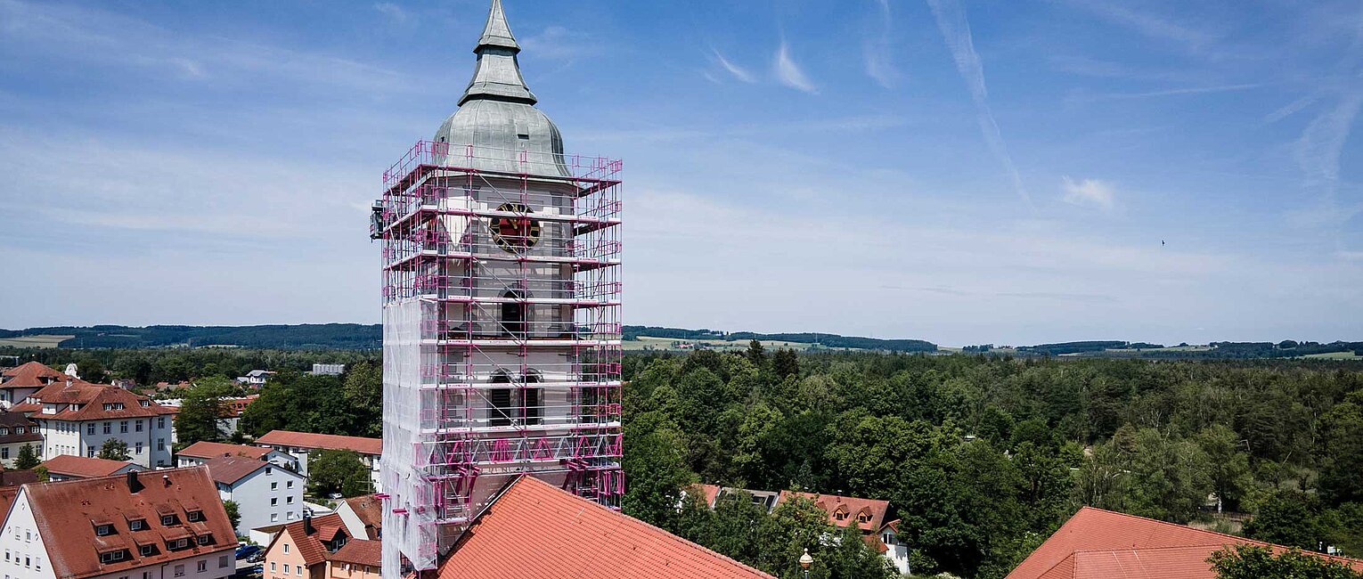Kirchturm mit Gerüst umgeben, rote Dächer im Vordergrund und grüne Landschaft im Hintergrund unter blauem Himmel.