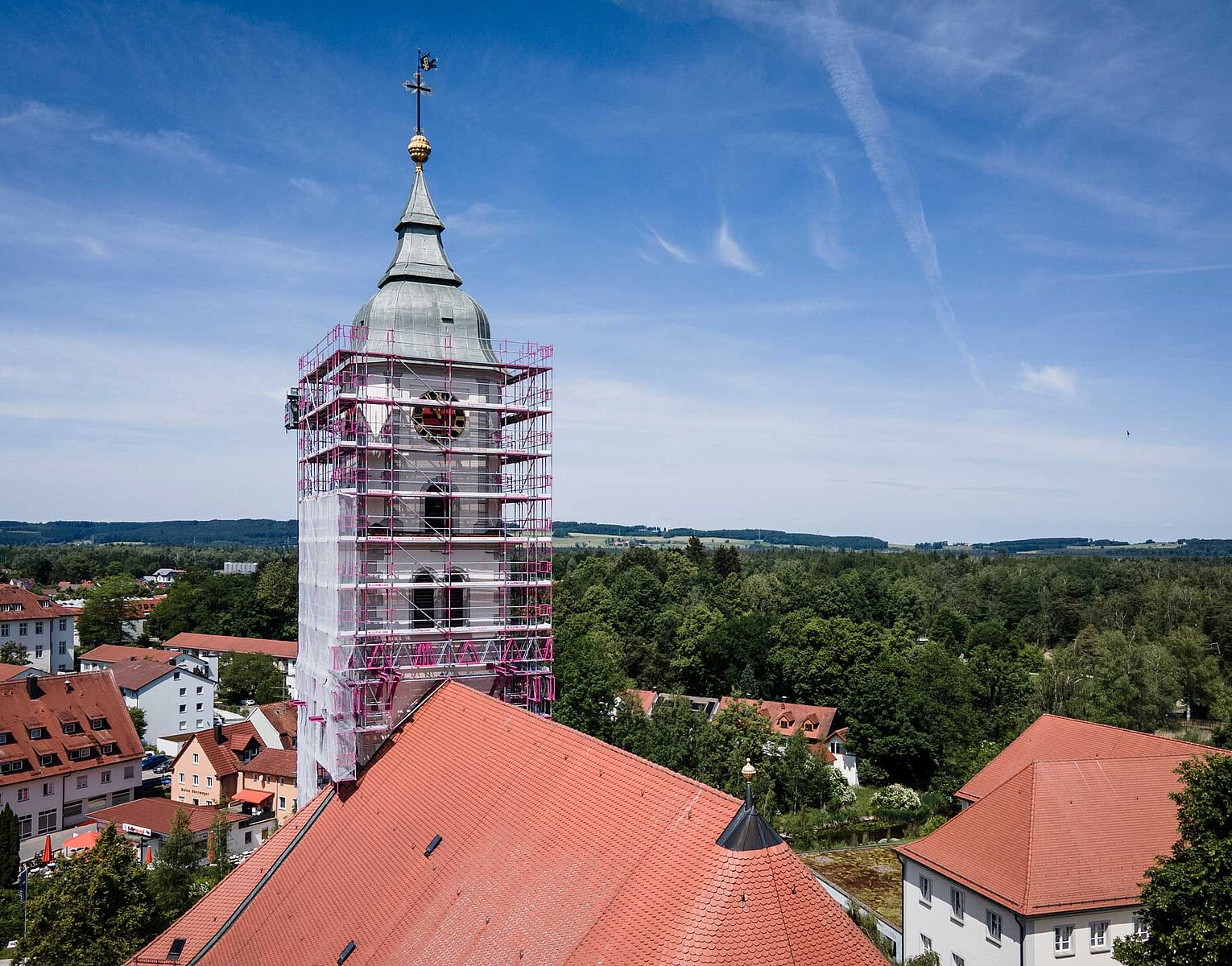 Kirchturm mit Gerüst umgeben, rote Dächer im Vordergrund und grüne Landschaft im Hintergrund unter blauem Himmel.