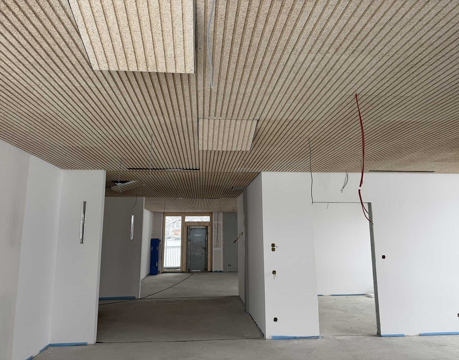 Innenraum eines Gebäudes mit weißen Wänden und einer Holzlamellendecke, der sich im Rohbau oder einer Renovierungsphase befindet. Einige Kabel hängen von der Decke, und der Boden ist unvollendet.