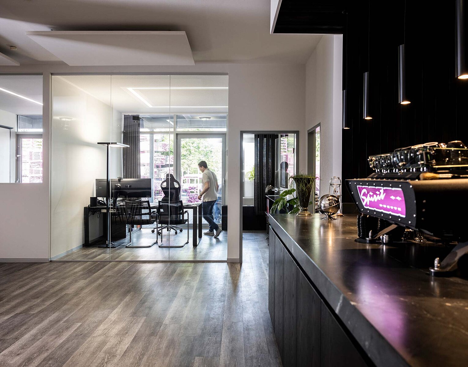 Gerüstbau Kaufmann Modernes Büro mit Glaswänden, einem Besprechungsraum, einem Schreibtisch, mehreren Stühlen und einer Person, die läuft. Im Vordergrund steht eine professionelle Espressomaschine auf einer dunklen Arbeitsplatte.