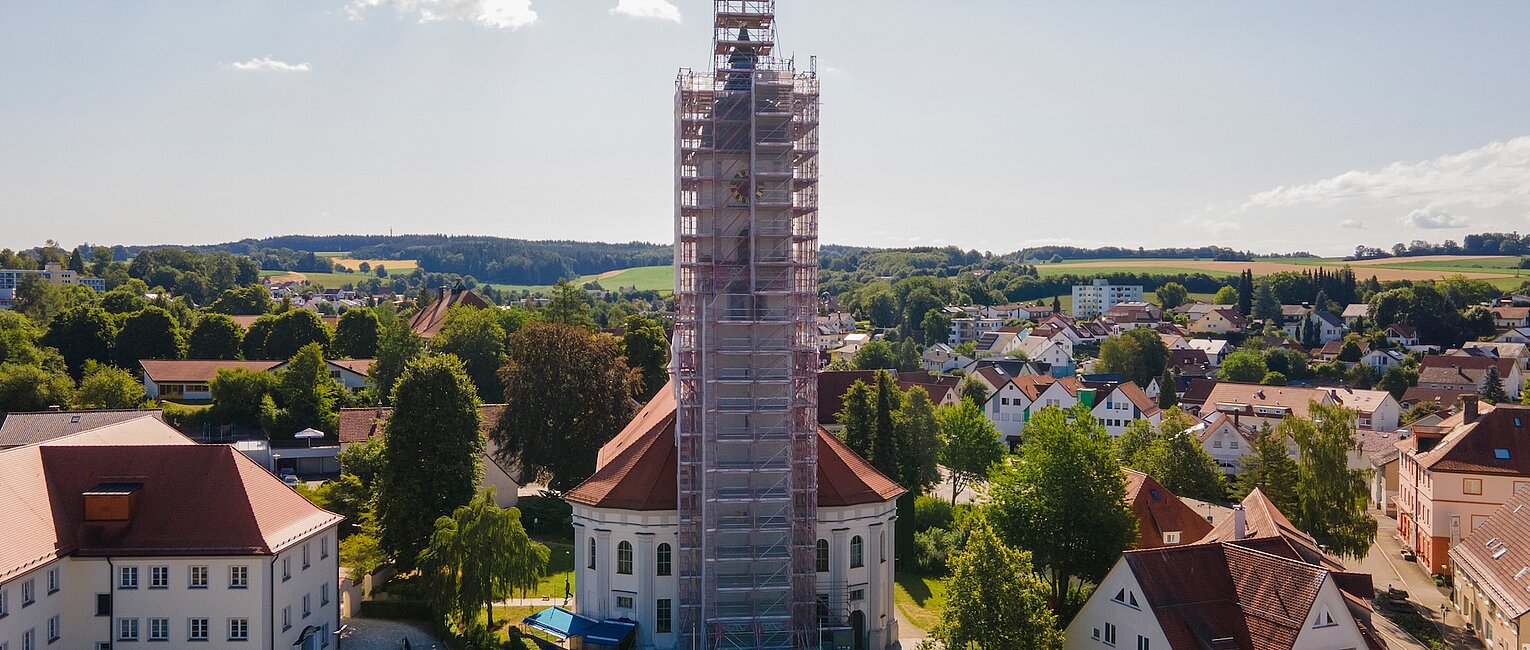 Blick auf ein rundes historisches Gebäude in einer Kleinstadt, das von Gerüsten umgeben ist, um Renovierungsarbeiten durchzuführen, umgeben von Wohnhäusern und grüner Landschaft im Hintergrund.
