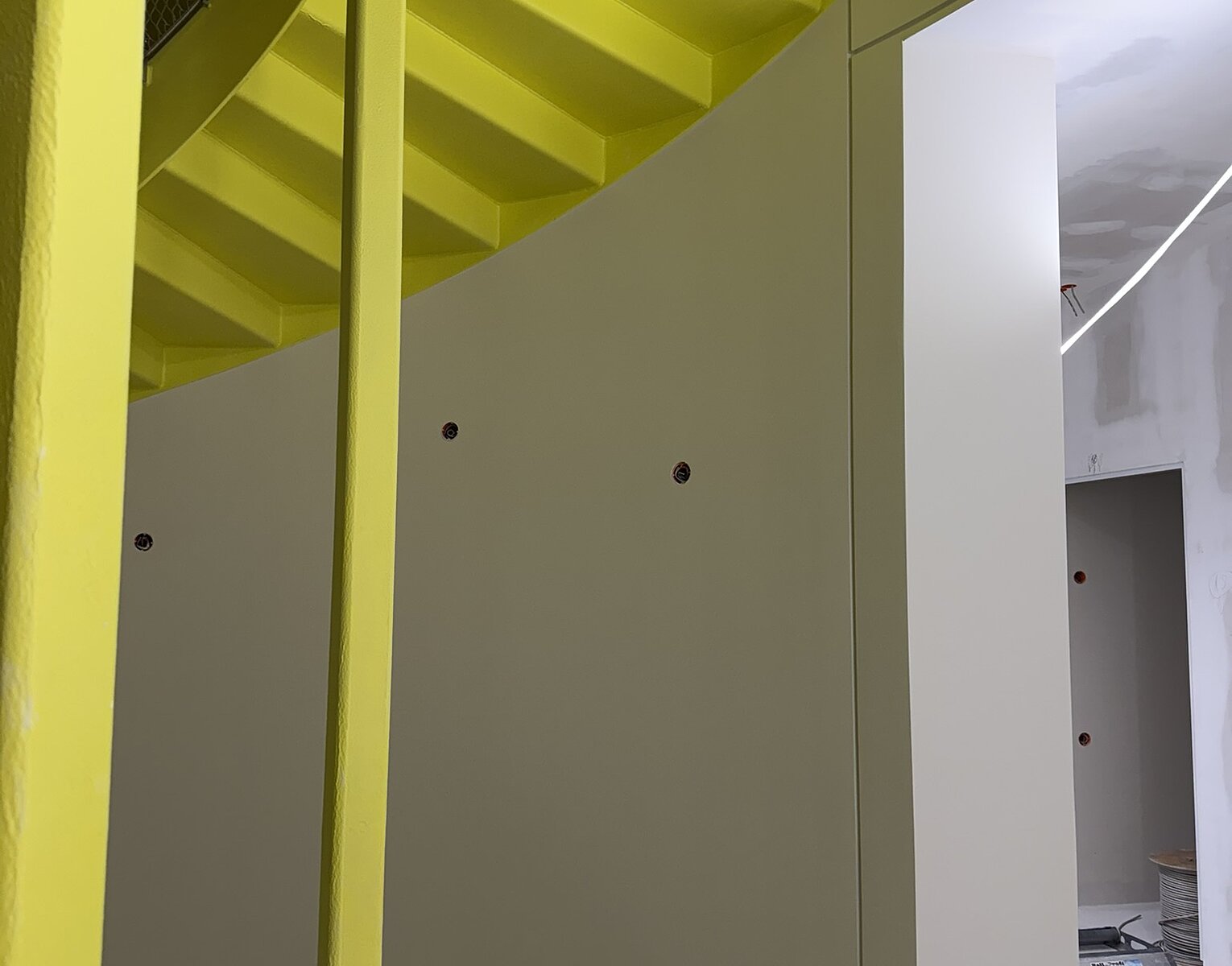 Gelbe Wendeltreppe in einem modernen Innenraum mit weißen Wänden und sichtbarem Baustellenbereich im Hintergrund.