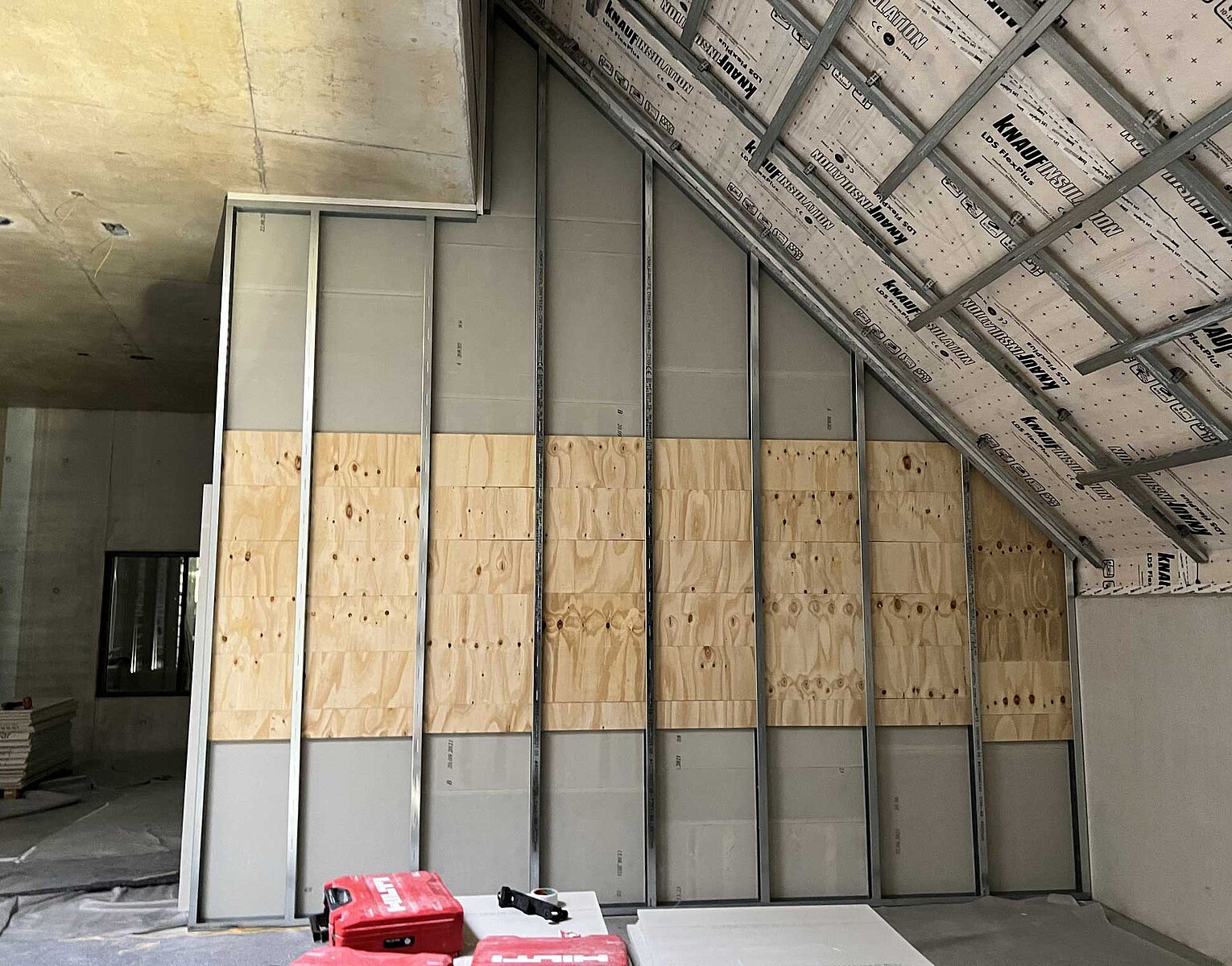 Unfertige Innenwandkonstruktion mit Metallrahmen und Teilschalung aus Holzplatten in einem Raum mit Dachschräge und sichtbarer Isolierung.