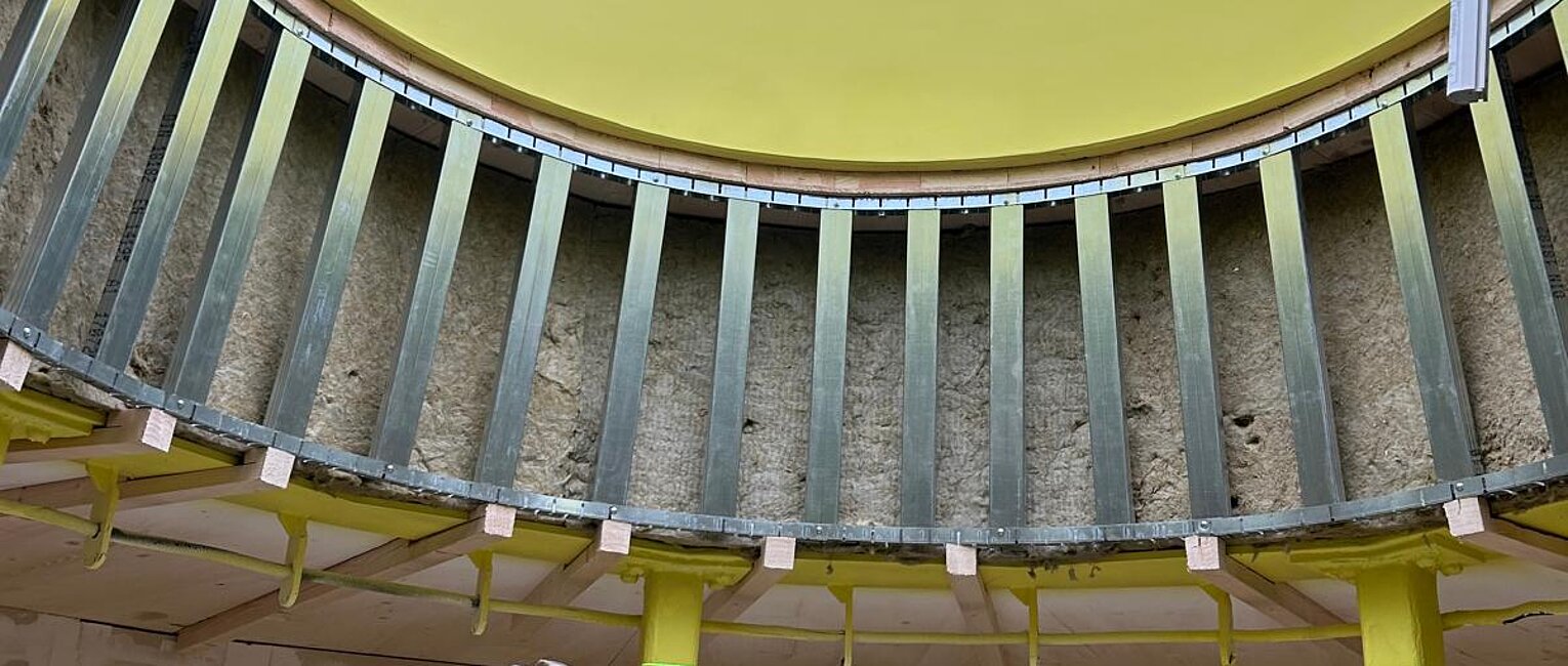 Blick in eine runde, gelb gestrichene Decke mit einem großen kreisförmigen Fenster, im Vordergrund steht eine Leitern, Bauarbeiten sind zu erkennen.