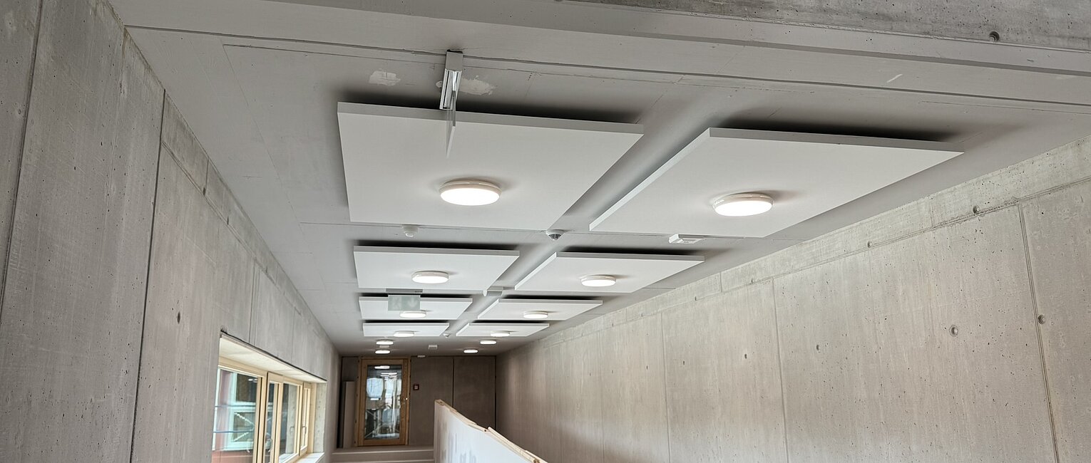 Blick in einen länglichen Flur mit rohen Betonwänden, Deckenlampen und einem Geländer, das eine Rampe abgrenzt. Rechts eine Treppe, links Fenster. Der Raum wirkt unfertig oder in Bauphase.