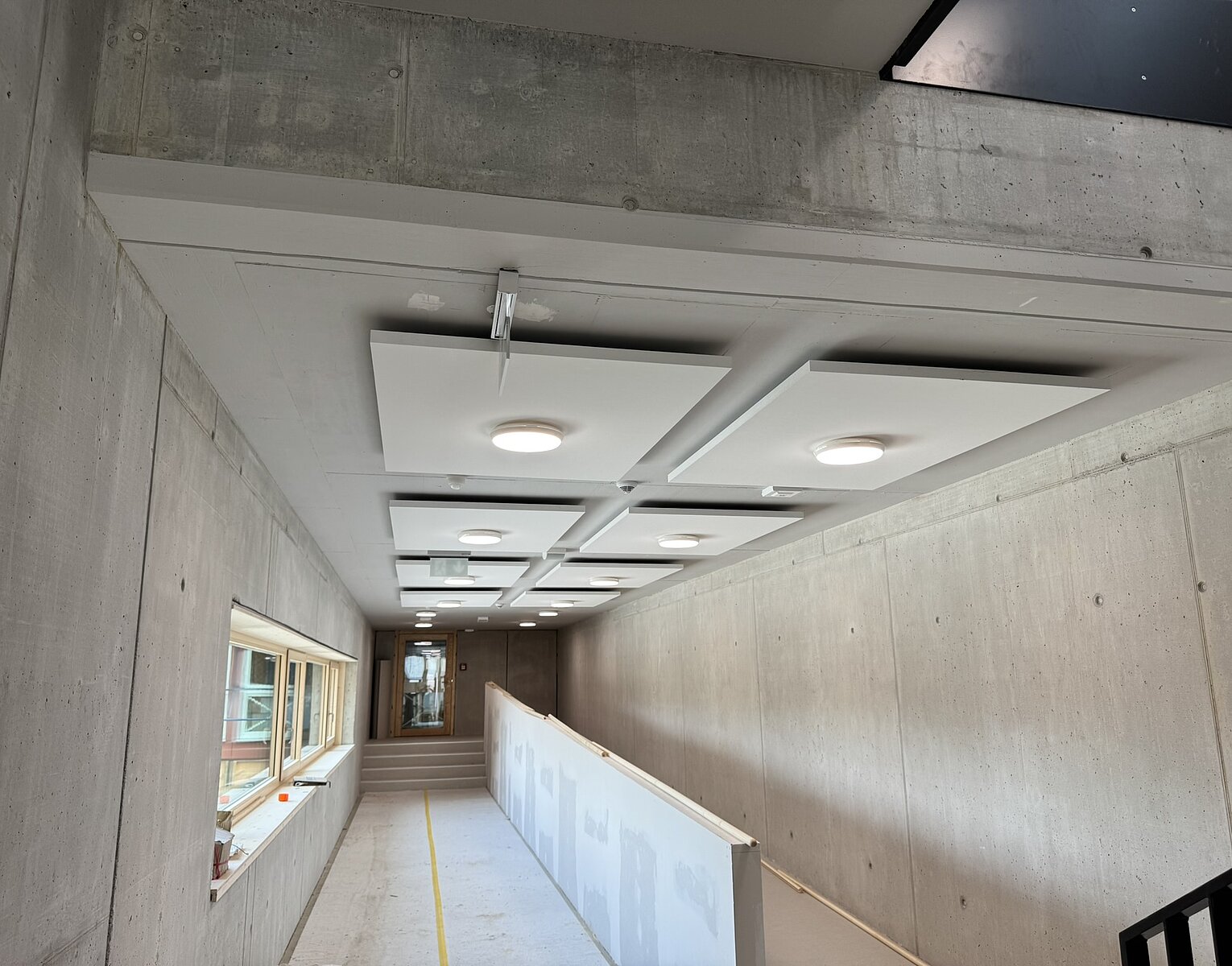 Blick in einen länglichen Flur mit rohen Betonwänden, Deckenlampen und einem Geländer, das eine Rampe abgrenzt. Rechts eine Treppe, links Fenster. Der Raum wirkt unfertig oder in Bauphase.
