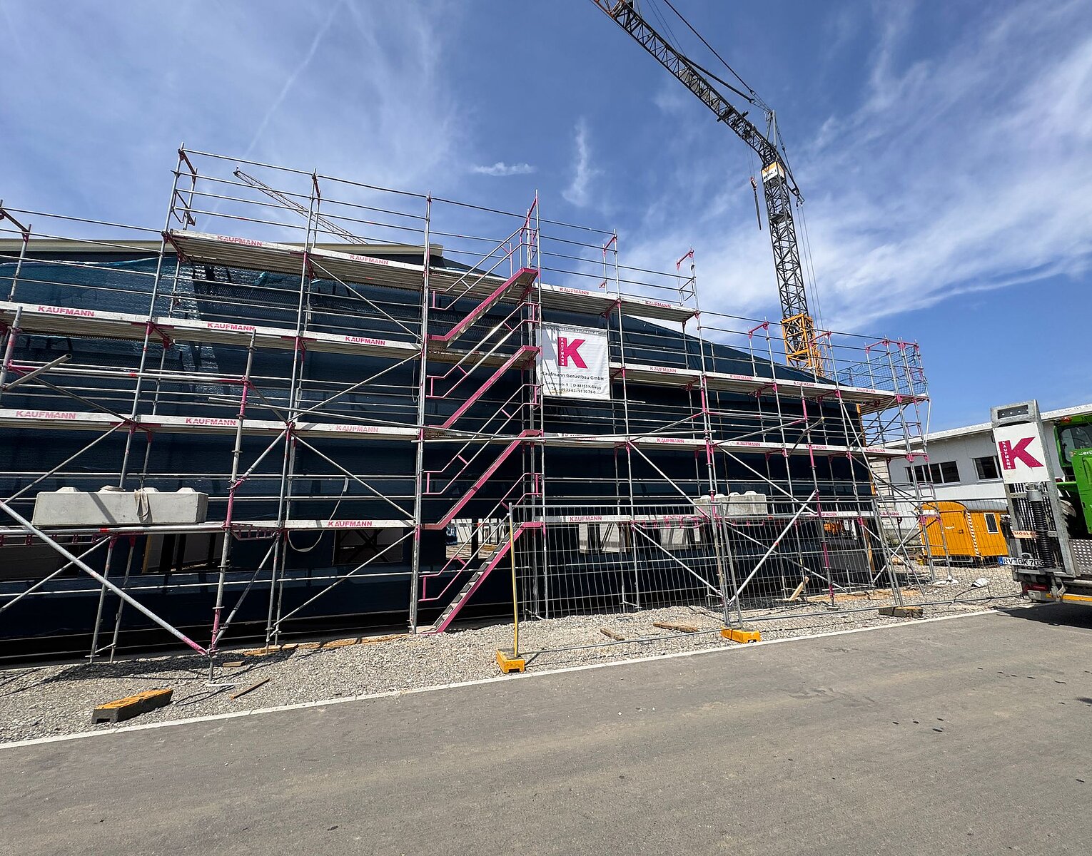 Baustelle mit Gerüst vor einem Gebäude, Kran und blauem Himmel im Hintergrund.