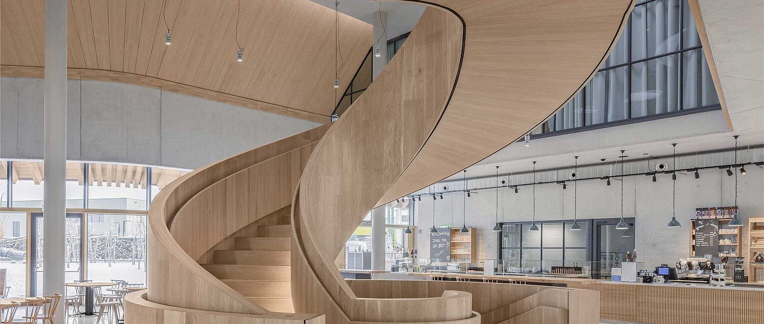 Innenaufnahme eines modernen Gebäudes mit einer spiralförmigen Treppe aus hellem Holz in der Mitte und großen Fenstern im Hintergrund.