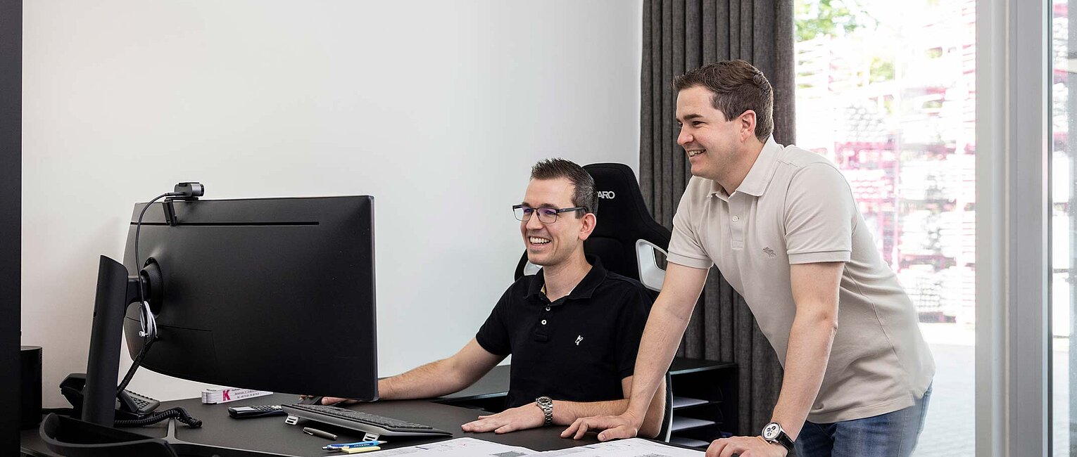 Zwei Männer arbeiten gemeinsam an einem Schreibtisch mit Computer und vielen Unterlagen in einem hellen Büro mit großen Fenstern.