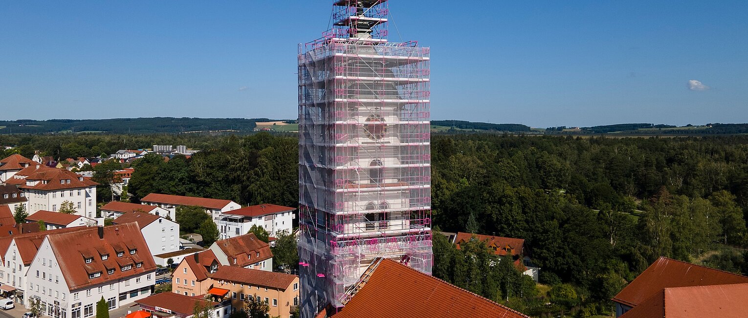 Kirchturm eines Gebäudes in einem Dorf, eingehüllt in ein Gerüst für Renovierungsarbeiten, umgeben von roten Dächern und viel Grün unter einem klaren blauen Himmel.