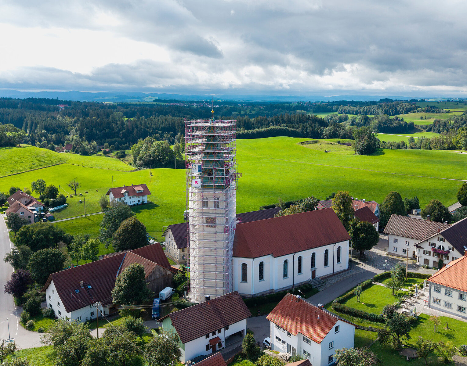 Luftaufnahme eines Dorfes mit einer Kirche, deren Turm mit Gerüsten eingerüstet ist, umgeben von grünen Feldern und bewaldeten Hügeln unter bewölktem Himmel.