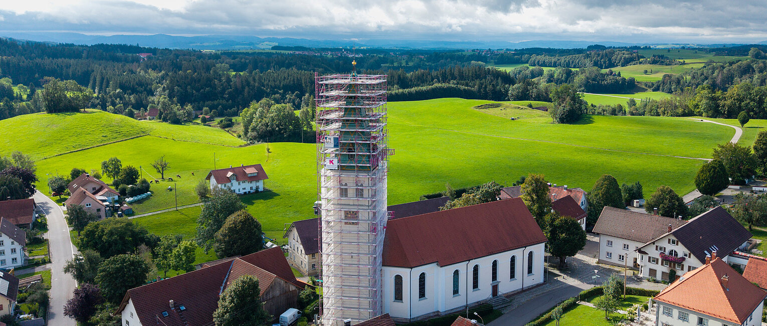 Luftaufnahme eines Dorfes mit einer Kirche, deren Turm mit Gerüsten eingerüstet ist, umgeben von grünen Feldern und bewaldeten Hügeln unter bewölktem Himmel.