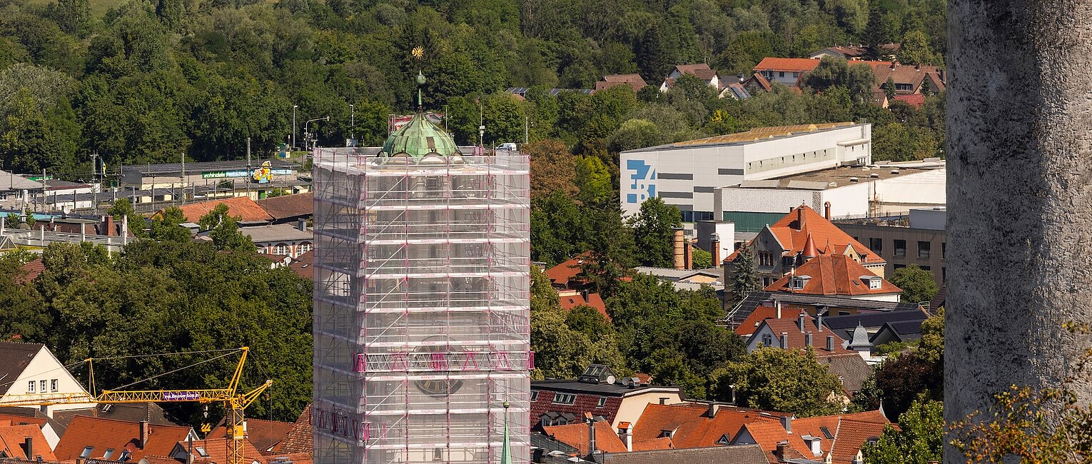 Stadtansicht mit Baustelle: Ein hoher Turm ist mit Gerüst und Schutznetz verhüllt, umgeben von roten Hausdächern und grünen Bäumen im Hintergrund.