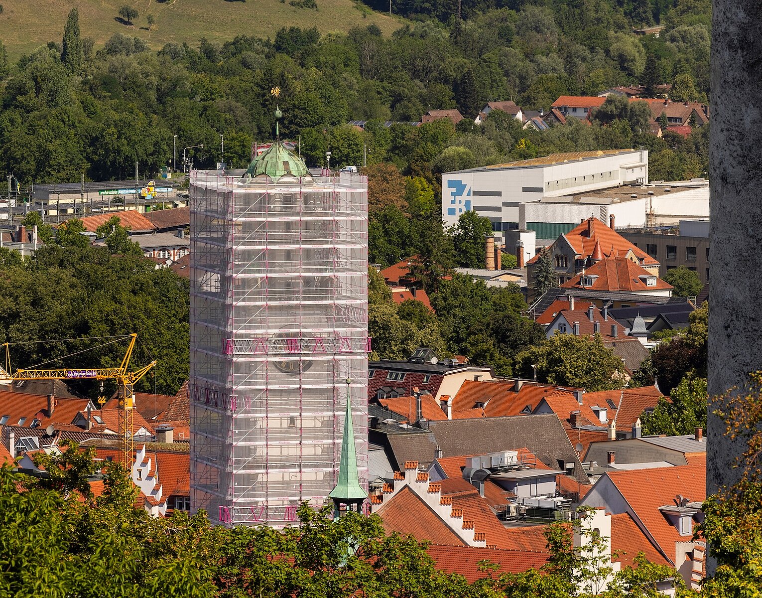 Stadtansicht mit Baustelle: Ein hoher Turm ist mit Gerüst und Schutznetz verhüllt, umgeben von roten Hausdächern und grünen Bäumen im Hintergrund.