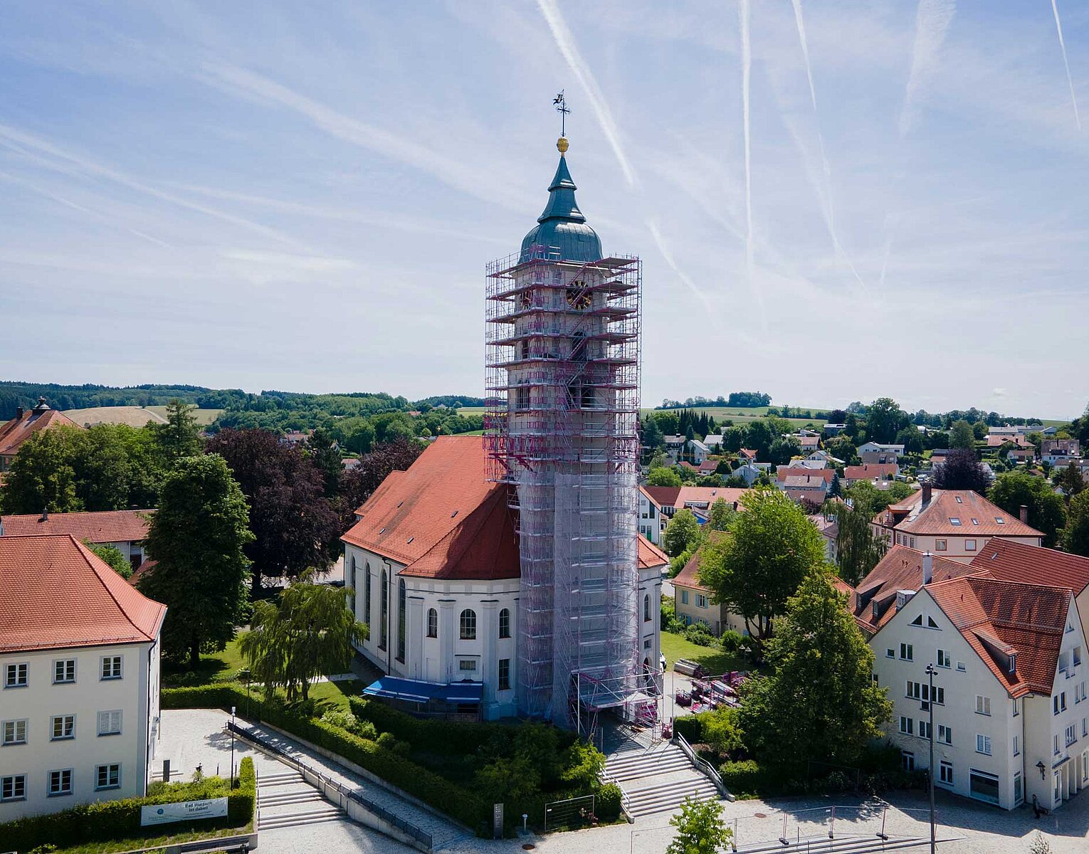 Kirchturm einer weißen Kirche mit rotem Dach, der von einem Gerüst umgeben ist, im Zentrum einer kleinen Stadt mit weiteren Wohnhäusern und grünen Bäumen unter einem blauen Himmel.