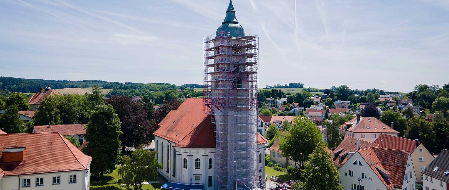 Kirchturm einer weißen Kirche mit rotem Dach, der von einem Gerüst umgeben ist, im Zentrum einer kleinen Stadt mit weiteren Wohnhäusern und grünen Bäumen unter einem blauen Himmel.