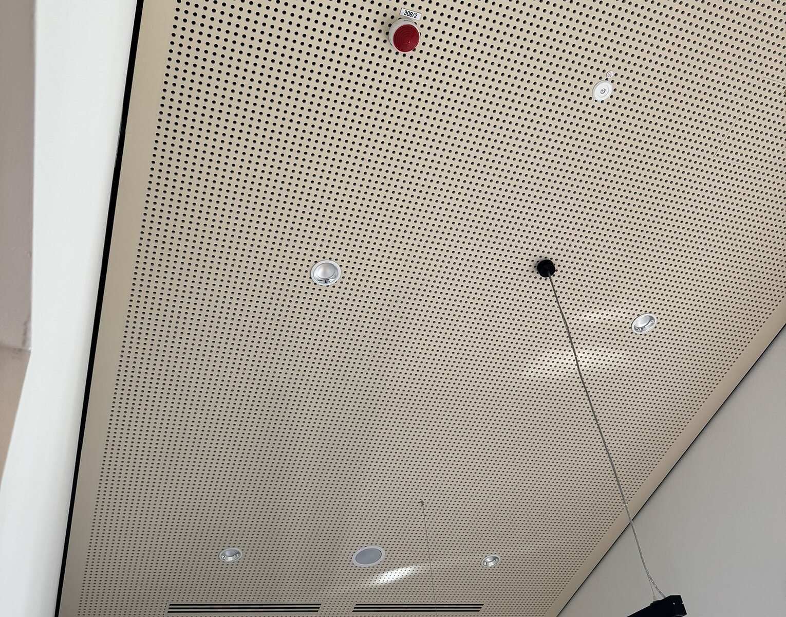 Decke mit Akustikplatten und integrierten Lampen, einem roten Rauchmelder und einer hängenden schwarzen Leuchte in einem modernen Innenraum.