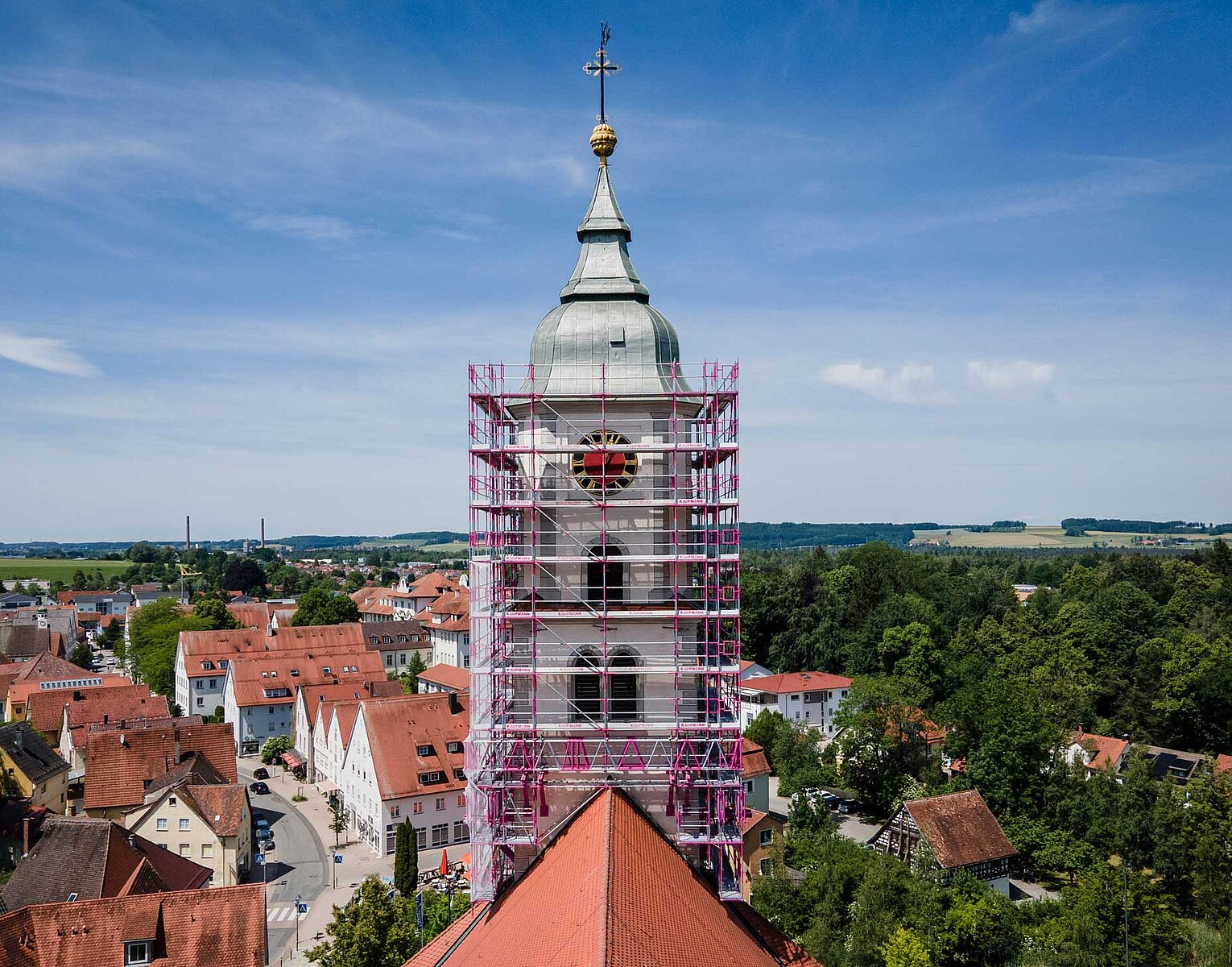 Kirchturm in einem Dorf, umgeben von Gerüst für Renovierungsarbeiten, mit roten Dächern und grünen Feldern im Hintergrund unter blauem Himmel.