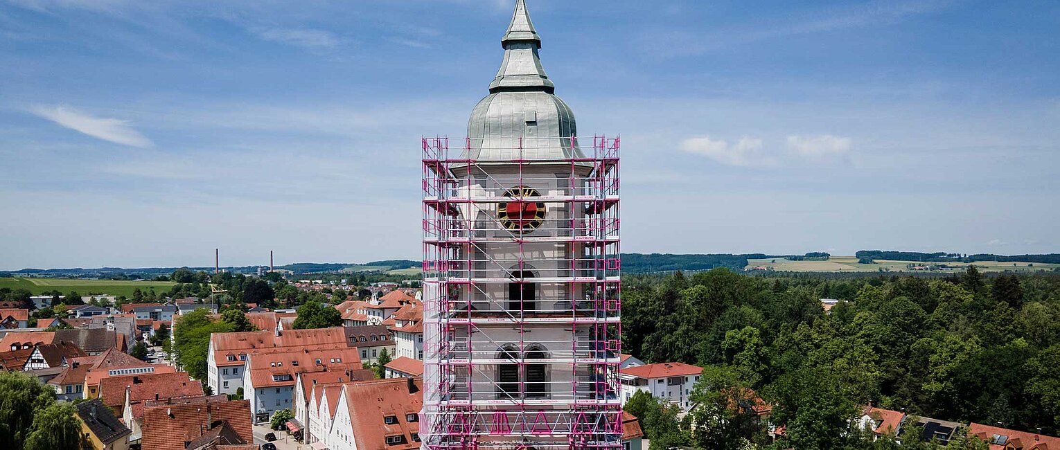 Kirchturm in einem Dorf, umgeben von Gerüst für Renovierungsarbeiten, mit roten Dächern und grünen Feldern im Hintergrund unter blauem Himmel.